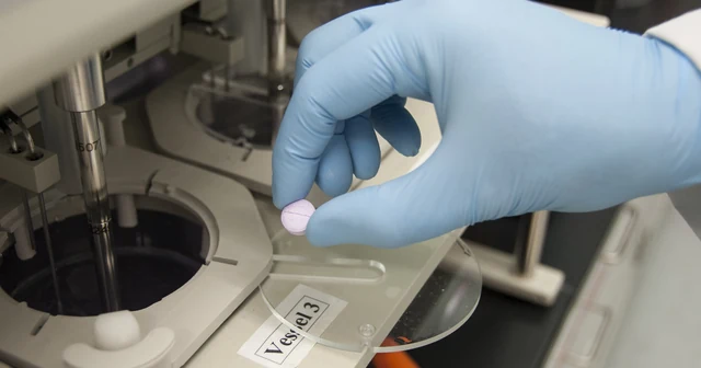 A gloved FDA scientist places a tablet into a dissolution testing vessel in a drug quality laboratory.